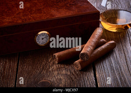 Stillleben mit Holzbox zur Befeuchtung, drei Zigarren und Glas Whiskey auf alten Tisch. Stockfoto