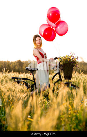 Hübsche Frau mit ihrem Fahrrad und mit roten Luftballons auf einer schönen Wiese Stockfoto