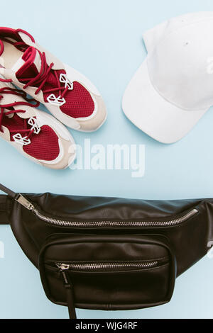 Sommer Layout der Kleidung eines jungen Mädchens. Modisch rot massive Sneakers, einem schwarzen Leder Gürteltasche und eine weiße Kappe auf einem Pastell-blaue Hintergrund. Stockfoto