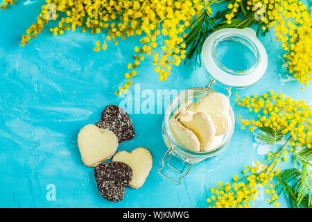 Köstliche gebackene Herzförmige Schokolade Cookies mit Kokos Chips in Glas und Silber wattle oder Mimosa auf blauen Beton Tabelle Oberfläche. Essen Stockfoto