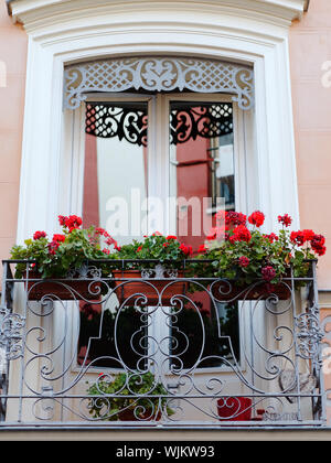Schöner Balkon mit weissen Fensterrahmen und roten Blüten im pflanzmaschinen hängen an Gusseisen dekorative Geländer. Stockfoto
