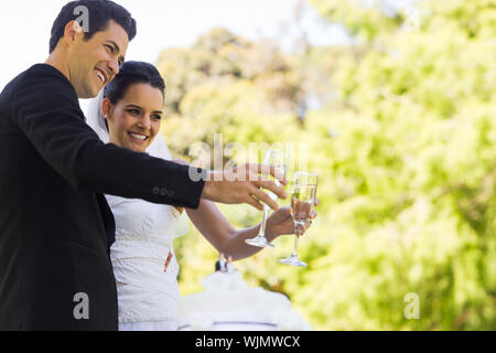 Glückliche junge jungvermählte Paar toasten Champagnergläser neben Kuchen im Park Stockfoto