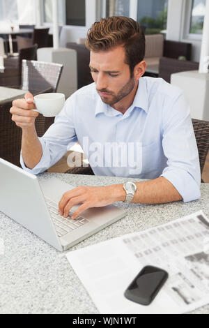 Stirnrunzelnd junge Unternehmer mit seinem Laptop holding Tasse Kaffee im Innenhof des Restaurants Stockfoto