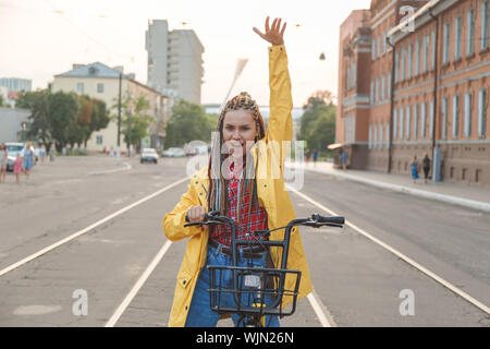 Portrait von hübschen Mädchen im gelben Mantel erhobenen Hand sitzen auf dem Fahrrad Stockfoto