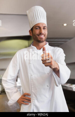 Fröhlicher junger Chef an Kameras mit Daumen hoch, die in einem kommerziellen Küche Stockfoto