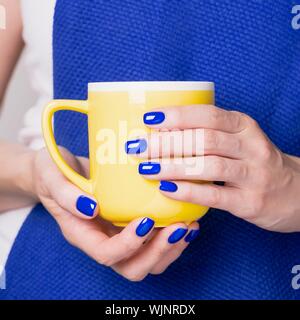 Schöne weibliche Hände halten gelbe Schale. Maniküre mit blauem Nagellack. Gemütliche Konzept Stockfoto