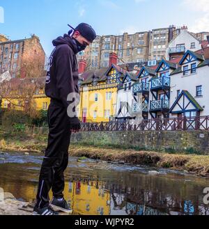 Eine Person, die einen Hoodie und eine schwarze Mütze trägt Ein schmutziger Teich in der Nähe von Gebäuden und Häusern Stockfoto