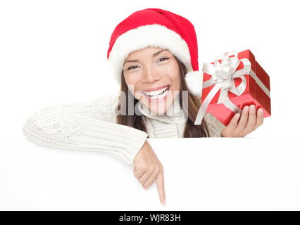 Weihnachten Frau beugte sich über Billboard unterzeichnen. Nach unten halten Geschenk zeigt große toothy Lächeln. Kaukasische/asiatische Frau mit Weihnachtsmütze und Winter Stockfoto