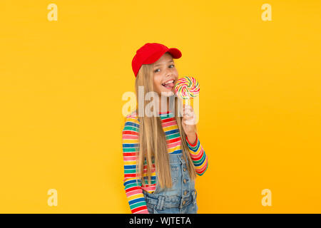 Lollipop macht Sie glücklich. Süße kleine Mädchen essen Regenbogen swirl Candy auf gelben Hintergrund. Kleines Kind lecken Hard Candy auf Stick. Adorable glückliches Kind genießen Kandiszucker Dessert. Candy Shop. Stockfoto