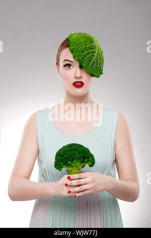 Porträt einer Frau zur Veranschaulichung einer Vegan-Konzepts mit einem Kohl auf dem Kopf und hält ein Brokkoli Stockfoto