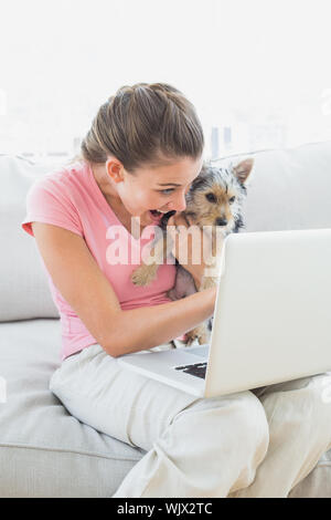 Gerne Frau ihren Yorkshire Terrier Holding auf die Couch mit Laptop zu Hause im Wohnzimmer Stockfoto