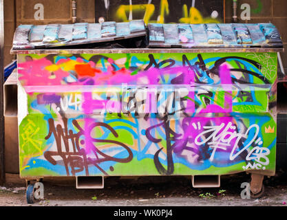 Ein Mülleimer oder Müllcontainer in einer Gasse voller Müll und abgedeckt im Graffiti Stockfoto