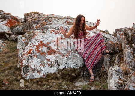 Freundliche hübsche Mädchen in einem rot gestreiften Kleid mit offenem Mund, Arme und Beine angehoben. Lehnt sich an ein großer Stein mit rotem Moos. Europäische Mädchen. Stockfoto