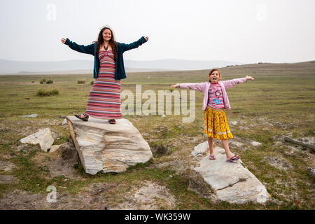 Fröhlich lächelnde Mutter und Tochter stehen auf große weiße Steine in der Steppe mit ausgestreckten Armen. Kind mit seiner Zunge heraus hängen. Stockfoto