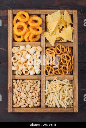 Verschiedene Snacks in Vintage hölzerne Kiste auf Holz- Hintergrund. Zwiebelringe, Nachos, salzige Erdnüsse mit Potato Sticks und Brezel. Geeignet für Bier und fi Stockfoto