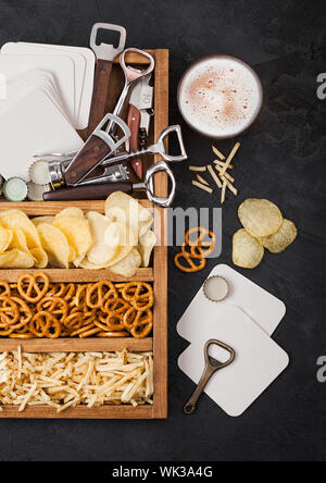 Glas Handwerk Lagerbier und Opener mit Box von Snacks auf schwarze Küche Tisch Hintergrund. Brezeln und Chips und salzig Kartoffelstifte in Vintage woode Stockfoto