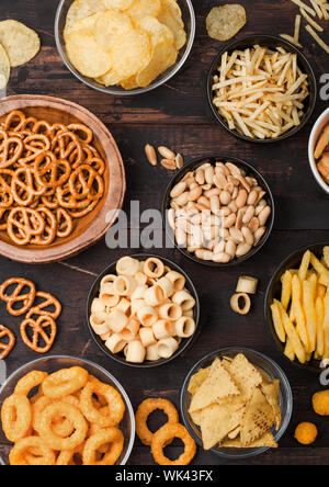 Alle klassischen Kartoffel Snacks mit Erdnüssen, Popcorn und Zwiebelringen und gesalzene Brezeln in Schüssel Platten auf Holz. Twirls mit Stöcken und Kartoffelchips und Cr Stockfoto