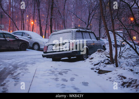 Autos im Schnee am Abend mit Spuren im Schnee geparkt. Straßenbeleuchtung Filterung durch die Bäume im Hintergrund. Stockfoto