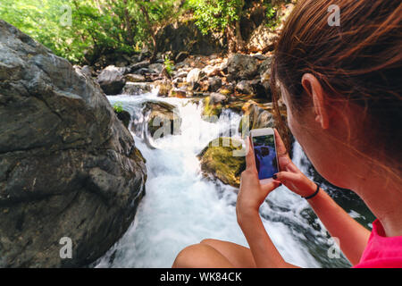 Bild einer jungen kaukasischen Frau mit roten Hemd von der Rückseite der Ihrer linken Schulter, während sie das Bild eines Wasserfalls mit Ihrem Telefon ist. Stockfoto