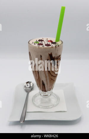 Schokoladenmilchshake mit dekorativer Schlagsahne und Schokoladenstreuer, serviert in einem hohen Glas mit grünem Stroh und Löffel auf einem weißen Teller. Stockfoto