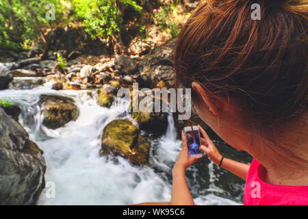 Junge kaukasier Frau mit roten T-Shirt mit dem Bild von einem Wasserfall mit Ihrem Telefon. Grosse Felsen und Bäume in der Umgebung der Kaskade. Stockfoto