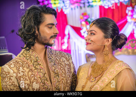 Indischer Bräutigam in Sherwani gekleidet mit herrlichem Braut stehend mit glamourösen Outfit und Schmuck mit Make-up und jede Hand in einem Bankettsaal halten. Stockfoto