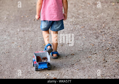 Rückansicht des unteren Abschnitt von einem 3-4 jährigen Kind im Sommer Kleidung auf einer Kiesgrube Fußweg Ziehen einer Spielzeug-LKW mit Kegel in Es Stockfoto