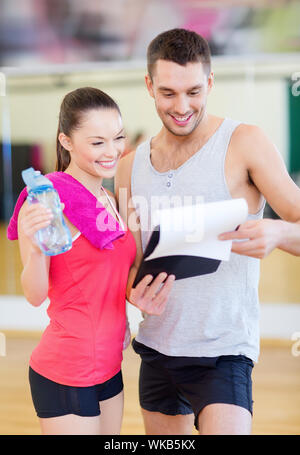 Fitness, Sport, Training, Fitness und Lifestyle Konzept - lächelnd männlichen Trainer mit Zwischenablage und Frau mit Wasserflasche in der Turnhalle Stockfoto