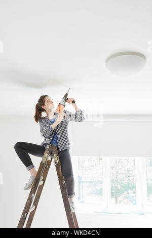 Die Frau als ein Handwerker auf einer Leiter mit einem Bohrer zum Bohren von Löchern in der Decke Stockfoto