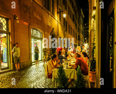 Nacht Blick auf die schmale Straße in Rom, Italien; speichert auf der linken und Outdoor Restaurant mit Menschen essen auf der rechten Seite Stockfoto