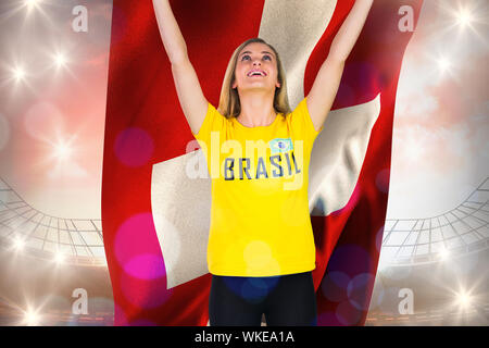 Aufgeregt Fußballfan in Brasil T-Shirt mit Schweizer Flagge gegen große Fußball-Stadion bei bewölktem Himmel Stockfoto