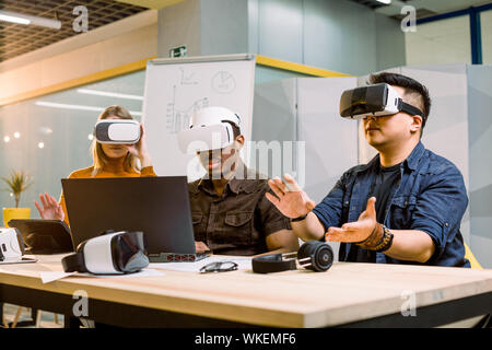 Junge Gruppe der multiethnical Menschen Spaß mit neuer Technologie VR-Headset. Kaukasische Mädchen, Afrikanische und chinesische Männer am Schreibtisch zu sitzen und zu versuchen, Stockfoto