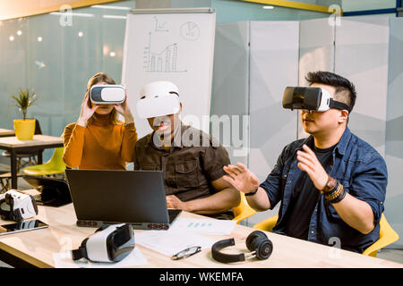 Junge Gruppe der multiethnical Menschen Spaß mit neuer Technologie VR-Headset. Kaukasische Mädchen, Afrikanische und chinesische Männer am Schreibtisch zu sitzen und zu versuchen, Stockfoto