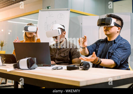 Junge Gruppe der multiethnical Menschen Spaß mit neuer Technologie VR-Headset. Kaukasische Mädchen, Afrikanische und chinesische Männer am Schreibtisch zu sitzen und zu versuchen, Stockfoto