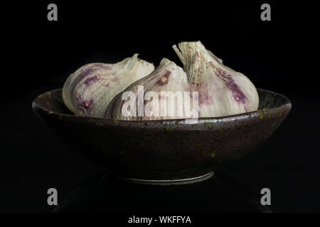 Gruppe von drei ganzen organische weiße Knoblauch allium sativum in dunklen Keramik Schüssel auf schwarzem Glas isoliert Stockfoto