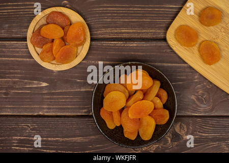 Menge ganze getrocknete orange apricot auf runden Bambus Untersetzer auf Bambus Schneidebrett in dunklen Keramik Schüssel flatlay auf braunem Holz Stockfoto