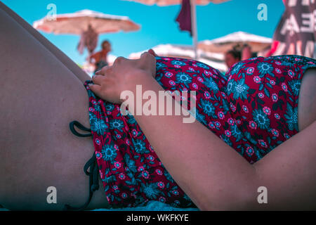 Schwangere Frau mit Händen auf den Bauch am Strand unter Dach ihr selbst zum Schutz vor der Sonne, Schwangerschaft und Sonne vermeiden Konzept Stockfoto