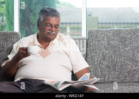 Asiatische indische Alter Mensch Kaffeetrinken beim Lesen der Zeitung Morgen, ältere Ruhestand innen leben Lebensstil auf Sofa zu Hause sitzen. Stockfoto