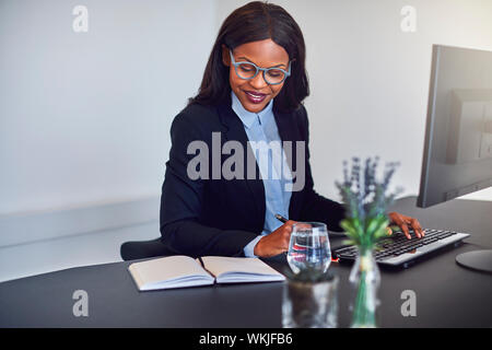 Lächelnden jungen afrikanischen amerikanischen Geschäftsfrau, die Computer und Lesen von Notizen, während an Ihrem Schreibtisch sitzen in einem Büro Stockfoto