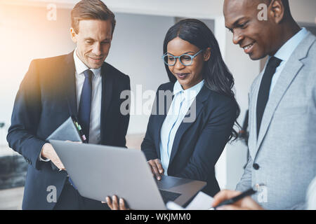 Vielfältige Gruppe von lächelnden Geschäftsleute stehen in einem hellen modernen Büro diskutieren gemeinsam auf einem Laptop Stockfoto