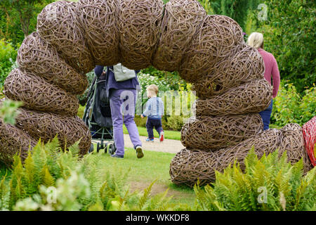 Kleinkind mit Mutter, die an einer großen, gewebten Weidenweide vorbeigeht, Caterpillar-Skulptur, RHS Garden, Harlow Carr, Harrogate, North Yorkshire, England, Großbritannien. Stockfoto