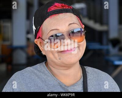 Malaysische chinesische Frau mit rot gefärbten Haaren trägt ein umgekehrt Baseball Cap und verspiegelten Sonnenbrillen und Lächeln für die Kamera. Stockfoto