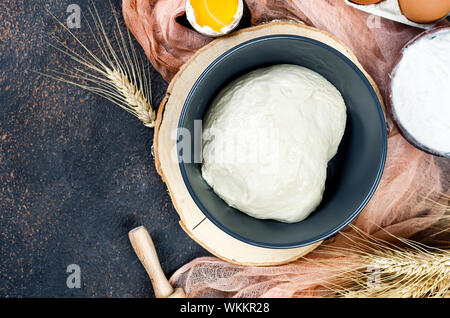 Rohe Hefe Teig in der Schüssel und Zutaten, Eier, Mehl, Ähren auf dunklen Küchentisch, Bäckerei Konzept, Stockfoto