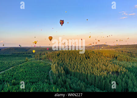 Bunte Heißluftballons in verschiedenen Formen fliegen über den Wald. Es ist ein Nadelwald im Sauerland. Die Sonne ist fast Einstellung, der Himmel ist b Stockfoto