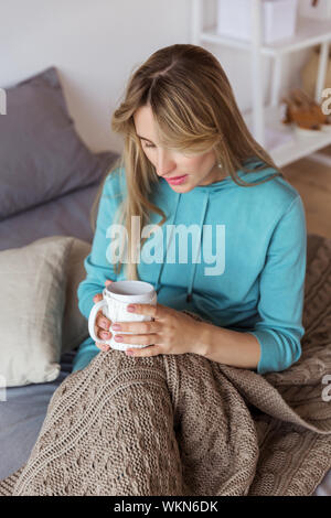 Ein Mädchen in einem blauen sweatshirt Stiche in eine Wolldecke und trinken heißen Tee verpackt Stockfoto