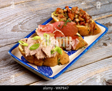 Anordnung der Tapas Bruschetta mit Pastete und Frühlingszwiebel, Speck, gebratenes Gemüse und Schinken auf Knoblauch Brot auf dem Teller auf weißem Holz- backgroun Stockfoto