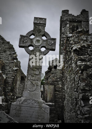 Grabstein mit keltischen Kreuz auf einem Friedhof mit Kirche Ruinen im Hintergrund Stockfoto