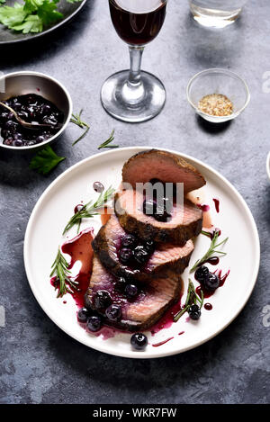 Roastbeef mit blaubeere Soße auf weiße Platte über blau Tisch aus Stein. Lecker medium gebratenes Roastbeef mit Berry Sauce. Stockfoto