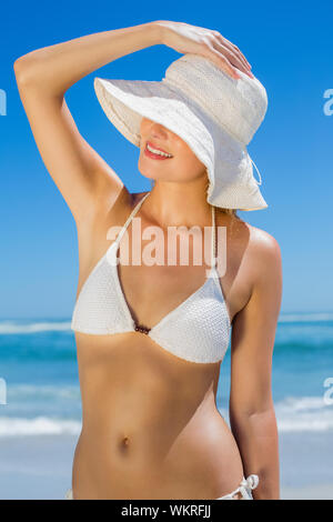 Lächelnde Blonde in white Bikini und Sonnenhut am Strand an einem sonnigen Tag Stockfoto