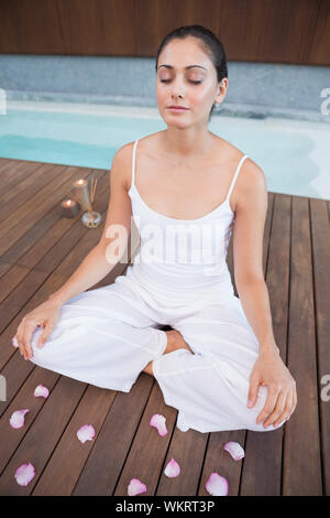 Friedliche Brünette in weißen Kleidern sitzen im Lotus Pose von Blütenblättern in Health Spa Umgeben Stockfoto